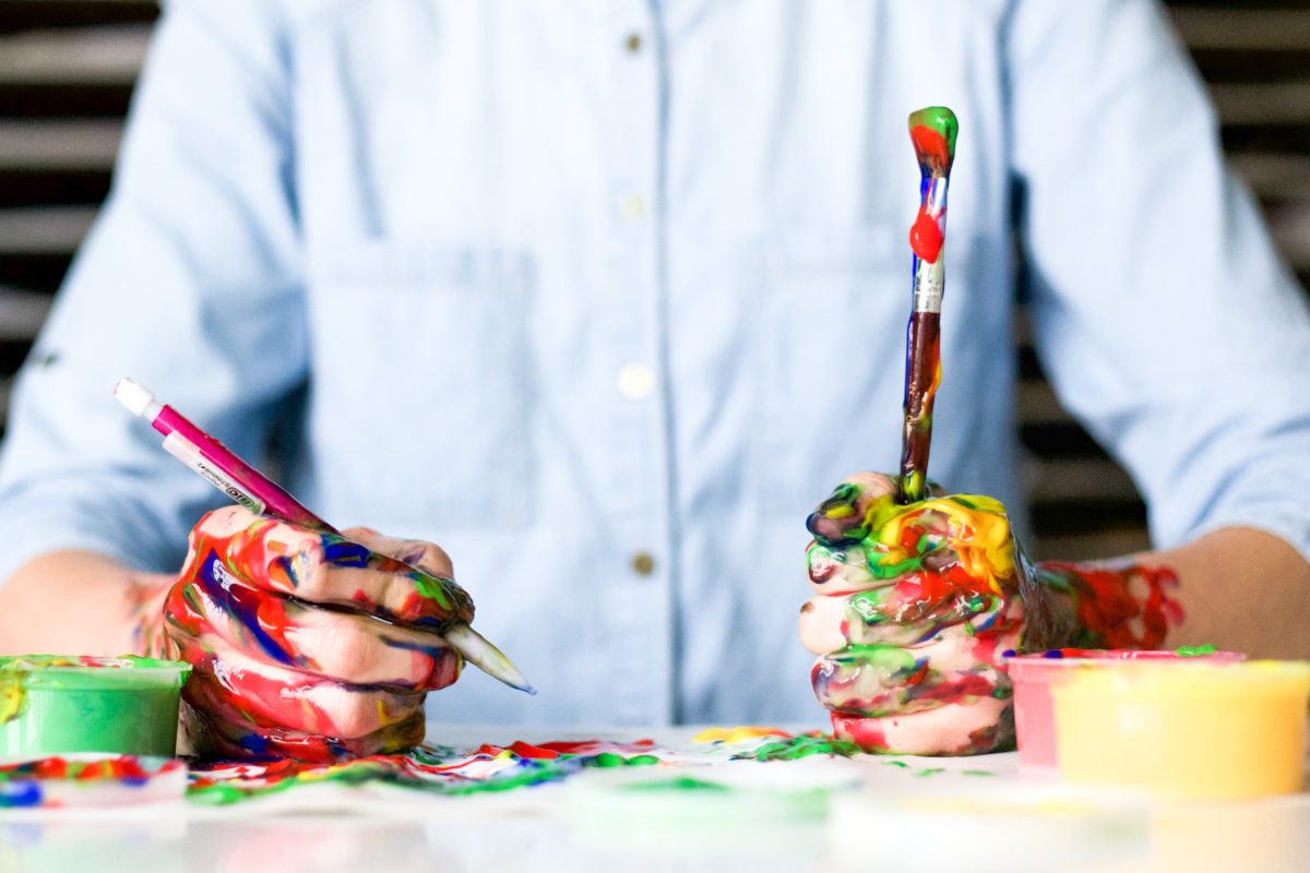 Torso shot of man with messy paint covered hands, holding two paint brushes.